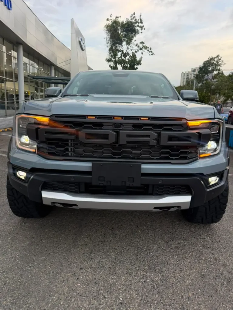 Vista frontal de una camioneta Ford Ranger Raptor gris con faros encendidos frente a un concesionario.
