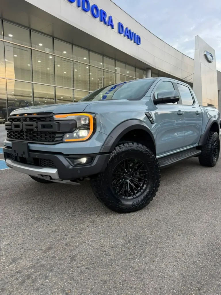 Camioneta Ford Ranger Raptor gris con llantas todoterreno frente a un concesionario.